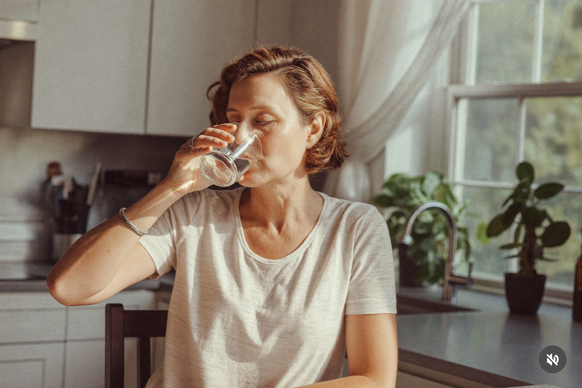 Woman in her fifties drinking a glass of water in a sunlit kitchen